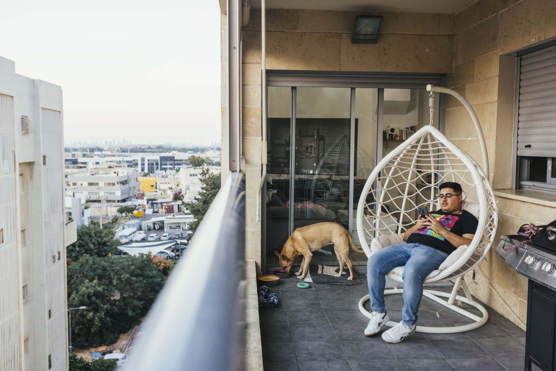  A man in dark T-shirt and jeans looks at his phone while seated in a white hammock chair on a balcony near a dog